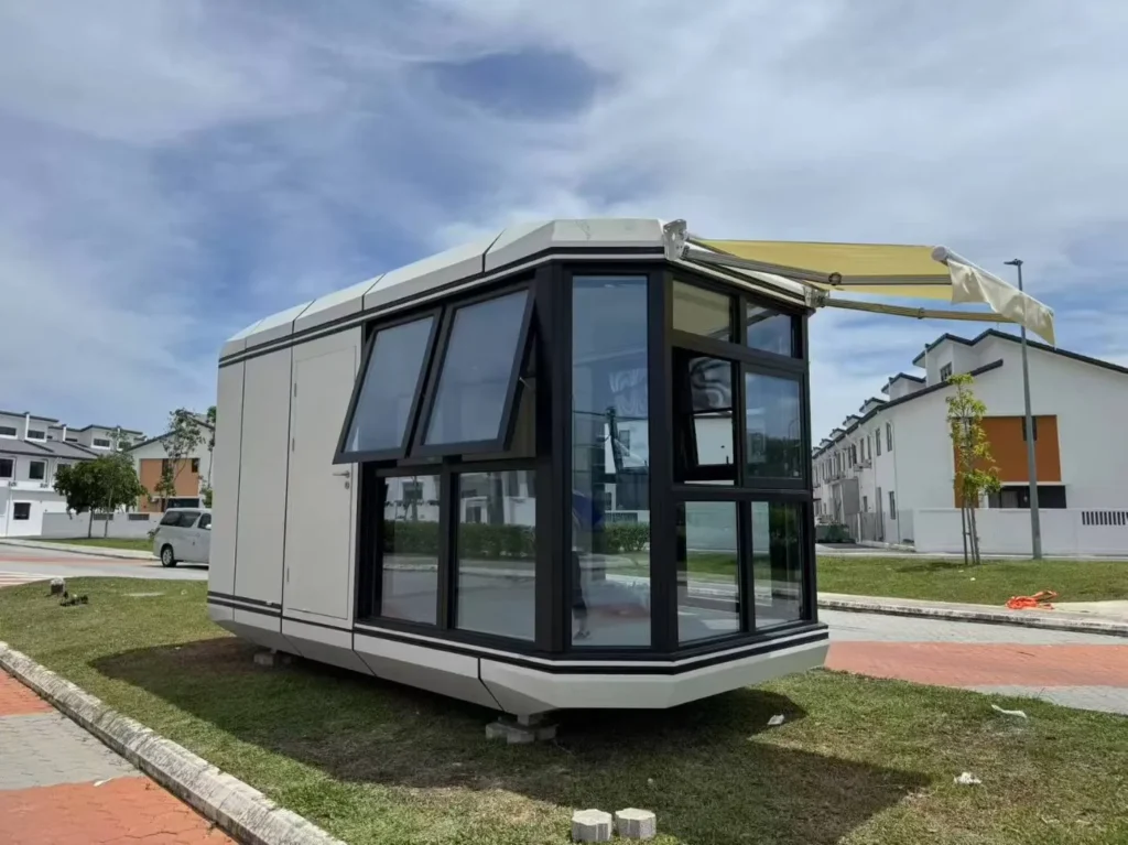 Light-coloured capsule with dark panoramic glazing, opening windows and an exterior awning outdoors