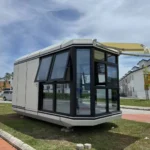 Light-coloured capsule with dark panoramic glazing, opening windows and an exterior awning outdoors