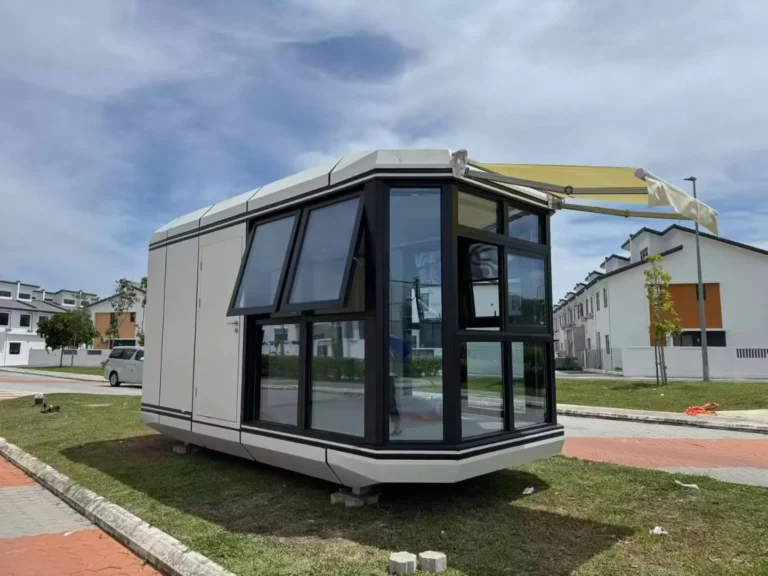 Light-coloured capsule with dark panoramic glazing, opening windows and an exterior awning outdoors