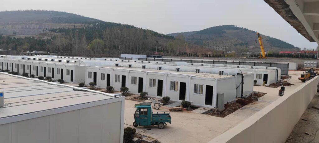 Large container house project site with rows of white modular units installed in a mountainous area, with construction equipment and a crane on site