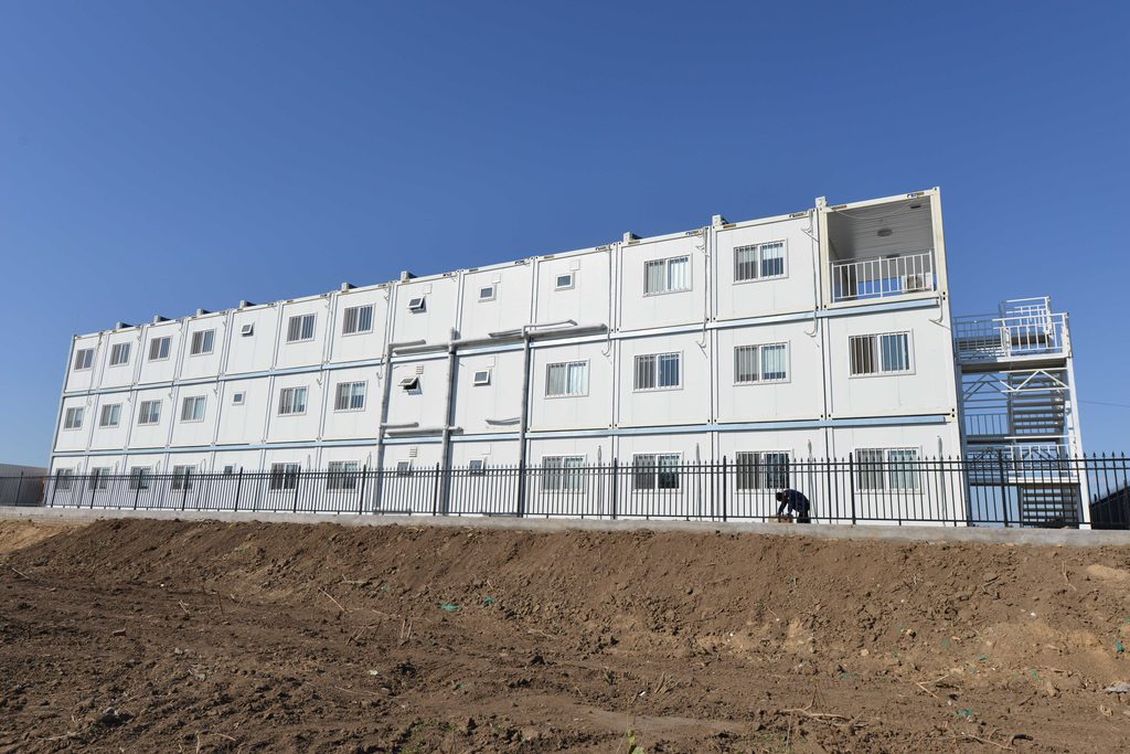 Multi-storey modular container house building with white panels and blue windows, standing on a construction site under a clear sky
